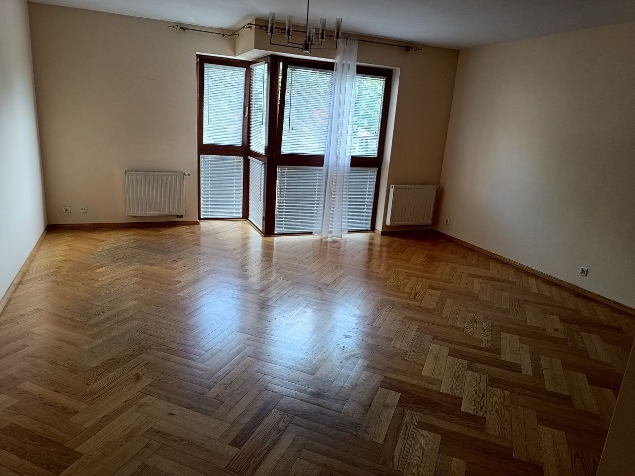 Pusty pokój z parkietem w jodełkę, kremowymi ścianami i dużym oknem z żaluzjami. Widoczne grzejniki i lampa sufitowa.