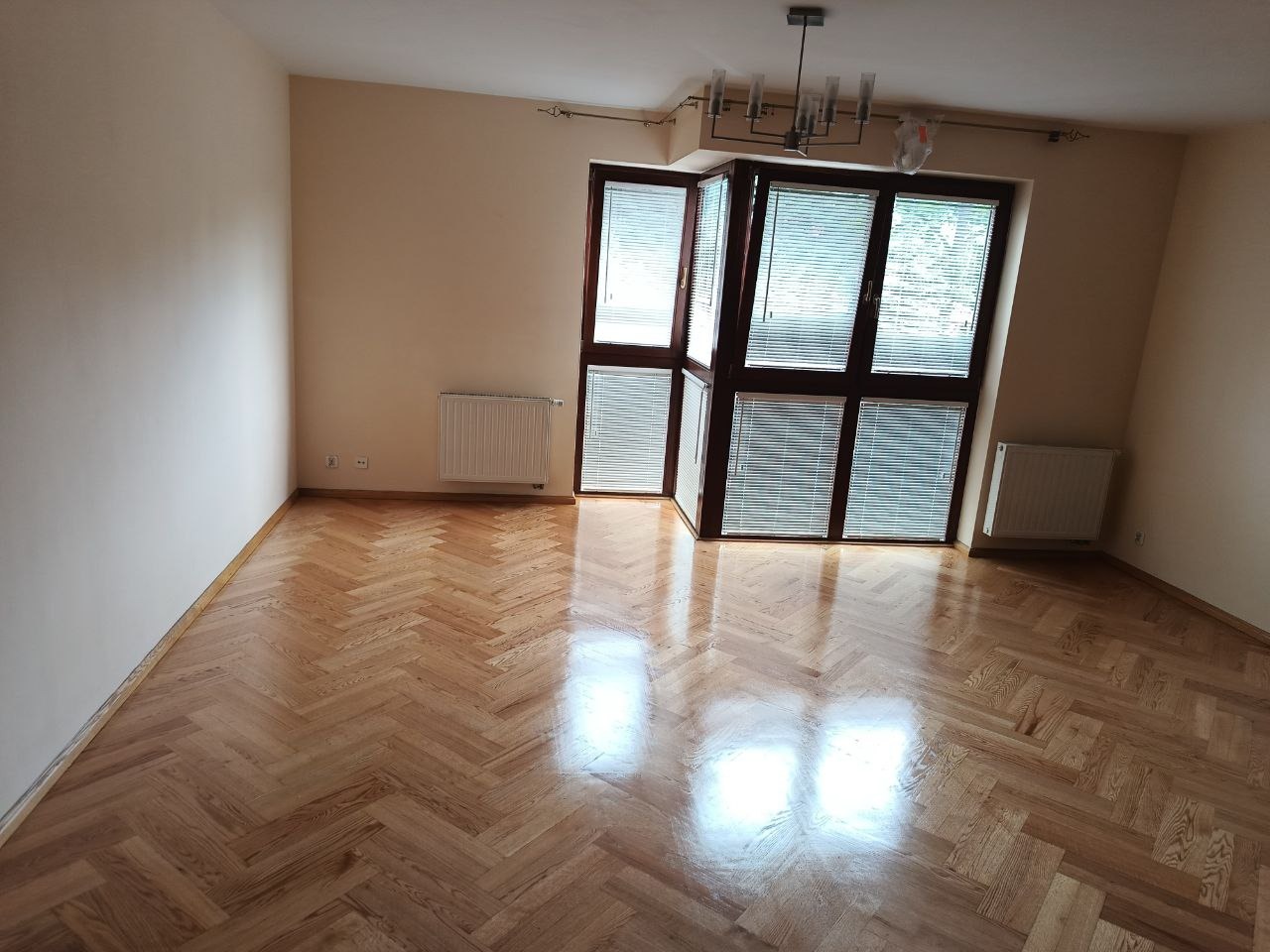 Jasny parkiet w jodełkę, świeżo położony w pustym pokoju z dużym oknem balkonowym i grzejnikami. Widok na wnętrze, ciepłe oświetlenie.