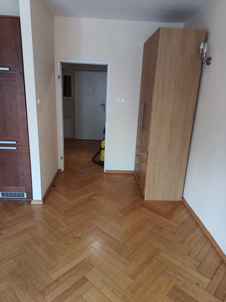 Parkiet ułożony w jodełkę, widok na szafę i otwarte drzwi. W głębi odkurzacz. Ciepłe oświetlenie.
