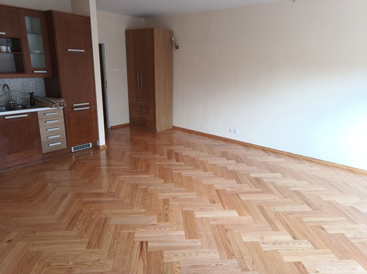 Jasny parkiet ułożony w jodełkę w pustym pokoju z brązowymi meblami kuchennymi i szafą. Świeżo wycyklinowana podłoga z widocznym połyskiem.