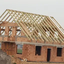 Surowy stan domu z cegły z zamontowaną drewnianą więźbą dachową, widok z poziomu gruntu na tle pochmurnego nieba i zabudowań gospodarczych w oddali.