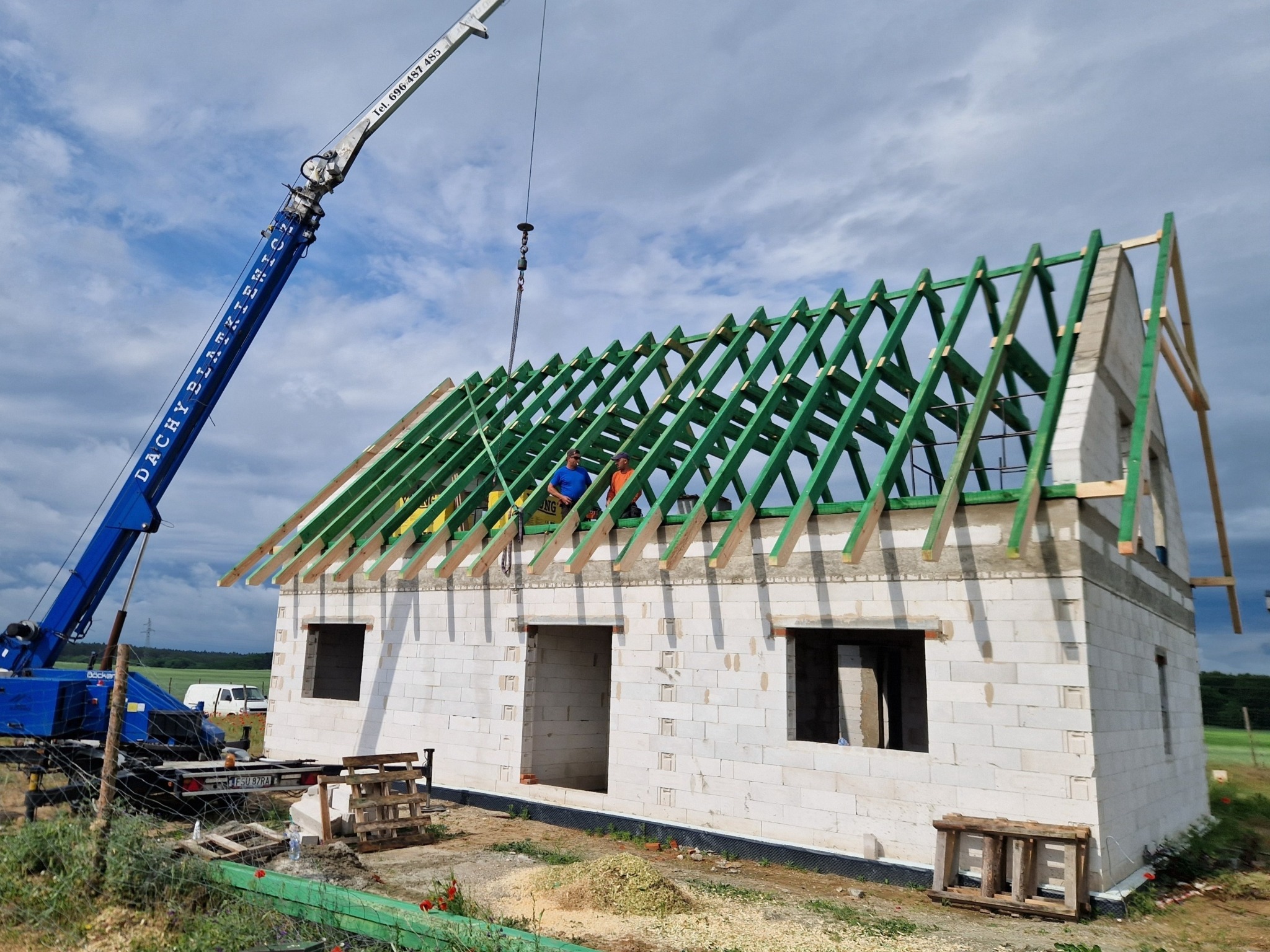 Montaż zielonej konstrukcji dachu na budynku z białego bloczka, widoczny dźwig z napisem 'Dachy Blachmix', pracownicy w pomarańczowych i niebieskich ubraniach na dachu.
