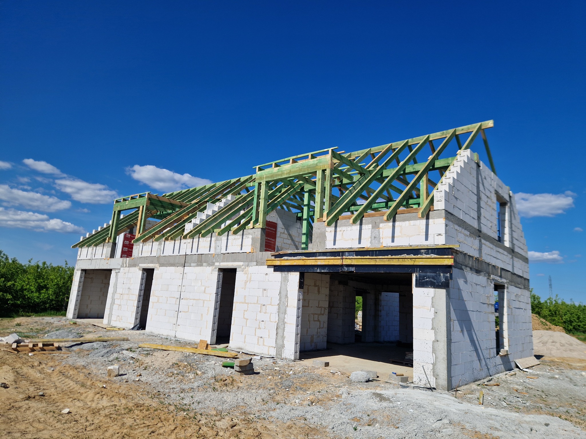 Surowy stan budynku z bloczków, z widoczną konstrukcją dachu z drewna, na tle błękitnego nieba z chmurami.