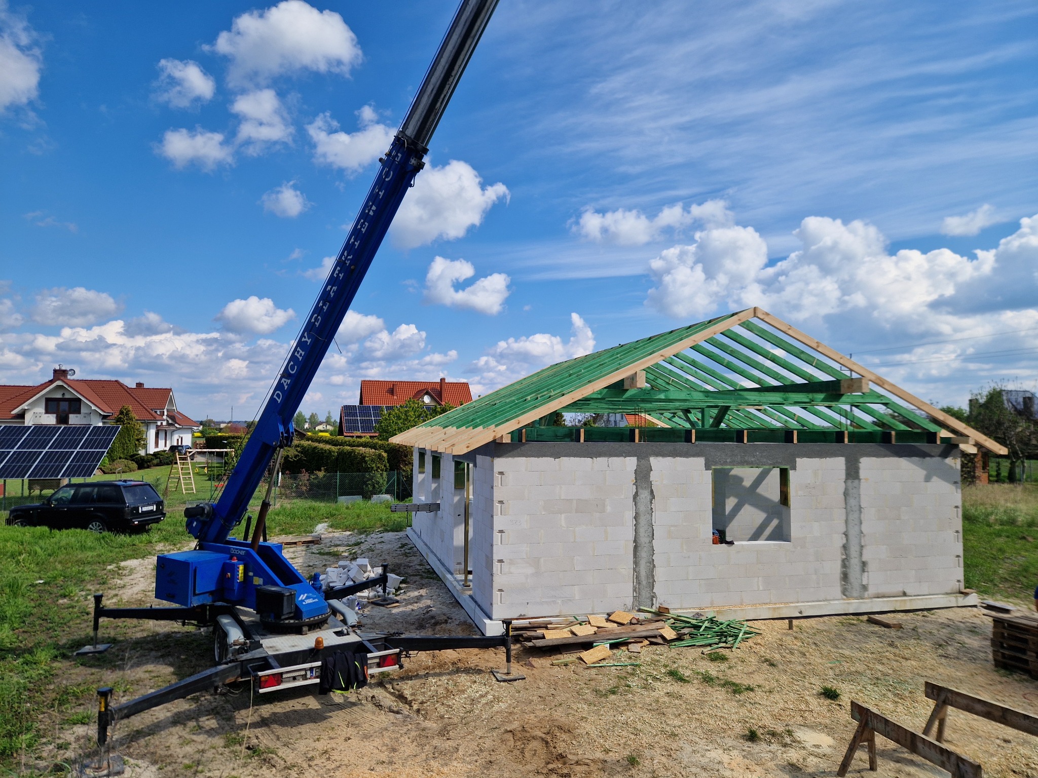 Budowa domu jednorodzinnego w stanie surowym otwartym, widoczny dźwig z wysięgnikiem i elementy konstrukcji dachu z zielonej impregnacji.