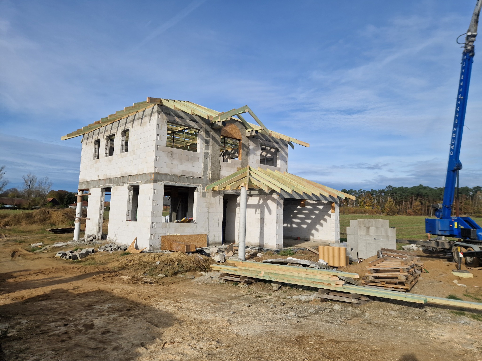 Wznoszenie więźby dachowej na dwukondygnacyjnym domu z bloczków betonowych, widoczny dźwig budowlany w tle.