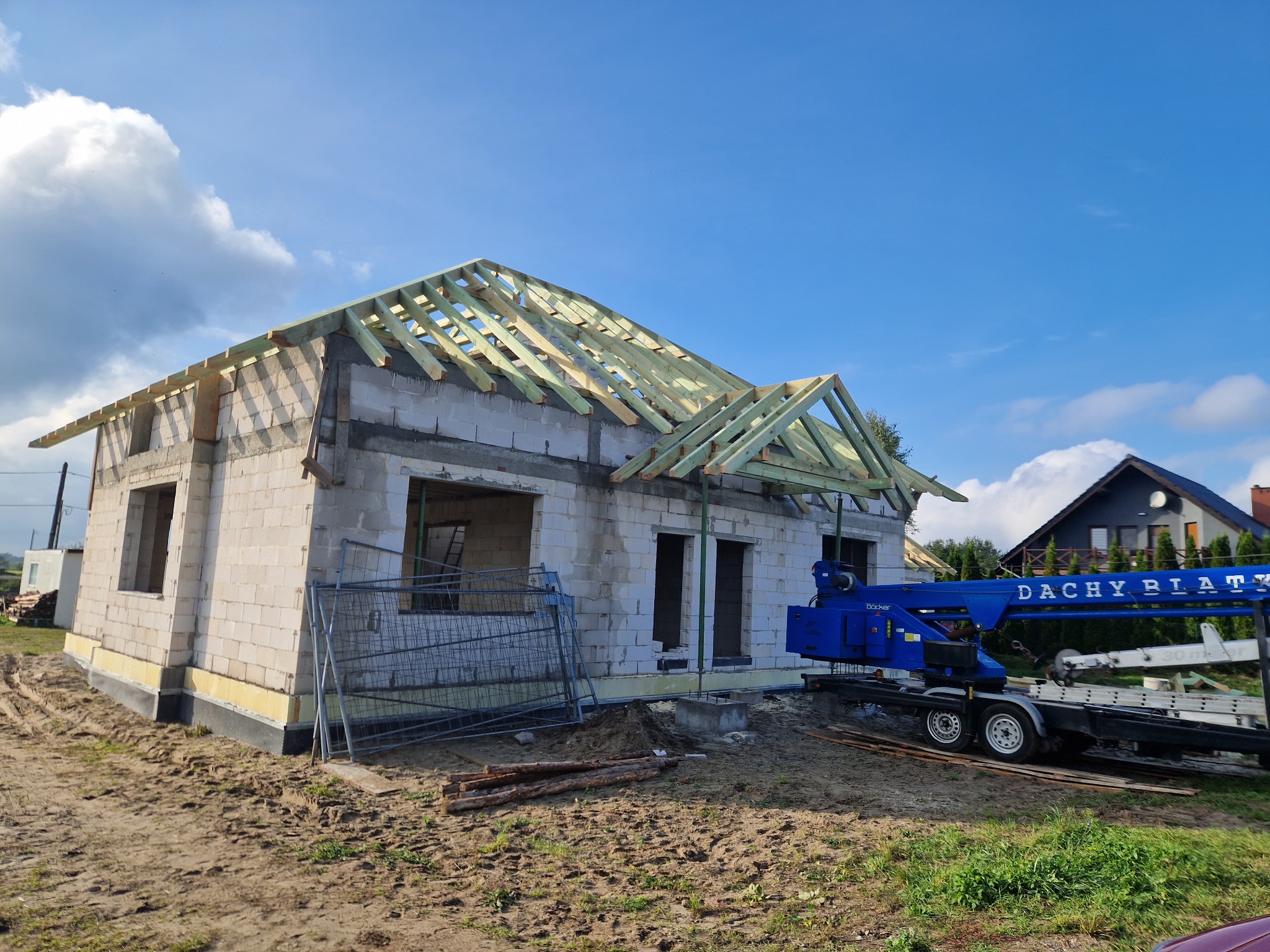 Budowa domu jednorodzinnego w stanie surowym otwartym, widoczna konstrukcja dachu z drewnianych belek, obok dźwig z napisem 'Dachy Piątek'.