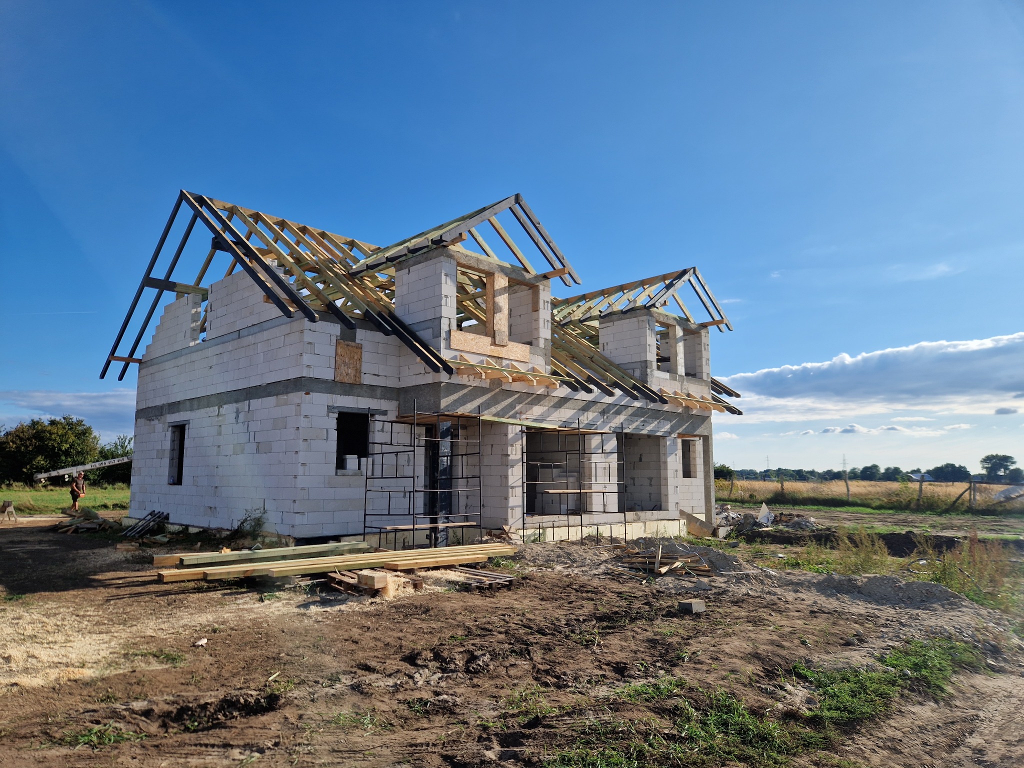 Surowy stan budynku z bloczków z częściowo zamontowaną konstrukcją dachu z drewnianych belek na tle błękitnego nieba i krajobrazu.