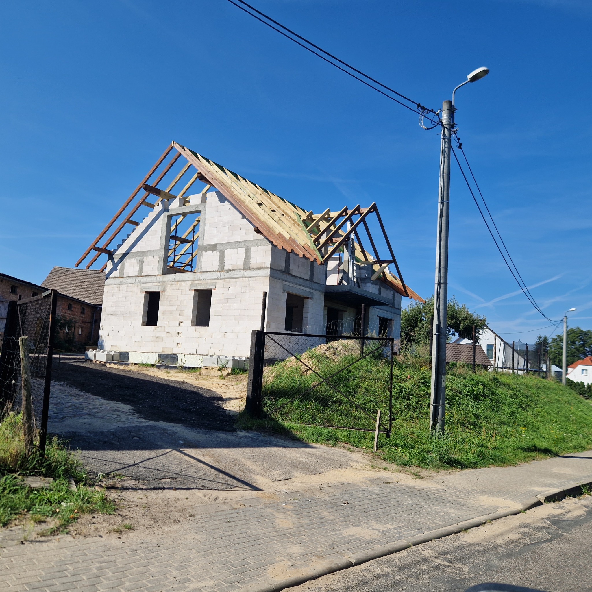 Budowa domu jednorodzinnego w stanie surowym otwartym z drewnianą konstrukcją dachu, widok z ulicy w słoneczny dzień.