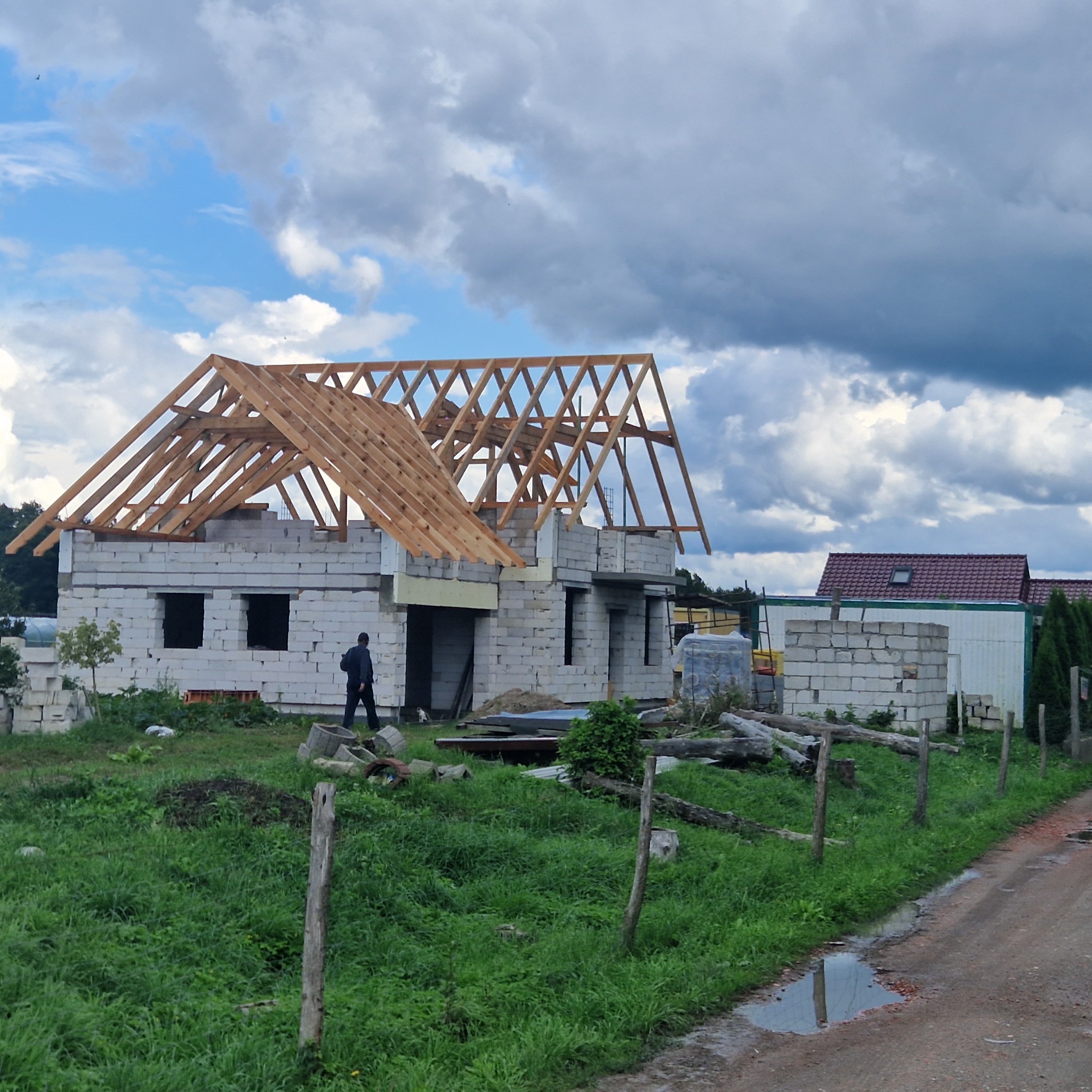 Budowa domu jednorodzinnego w trakcie realizacji: wzniesione mury z bloczków i drewniana konstrukcja dachu na tle pochmurnego nieba.