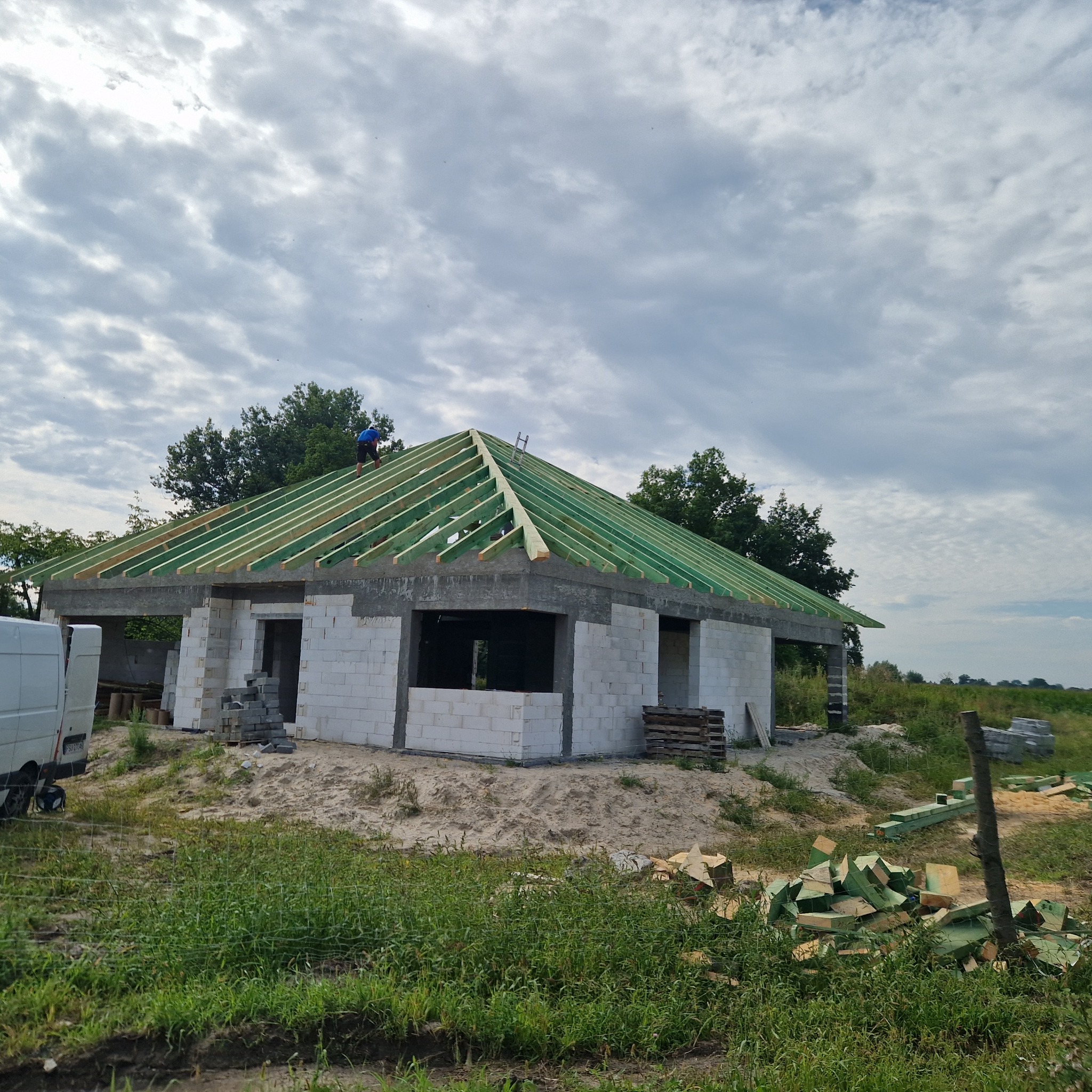 Wznoszenie drewnianej konstrukcji dachu na parterowym budynku z białego bloczka, widoczny pracownik na dachu i przygotowane elementy konstrukcyjne na ziemi.