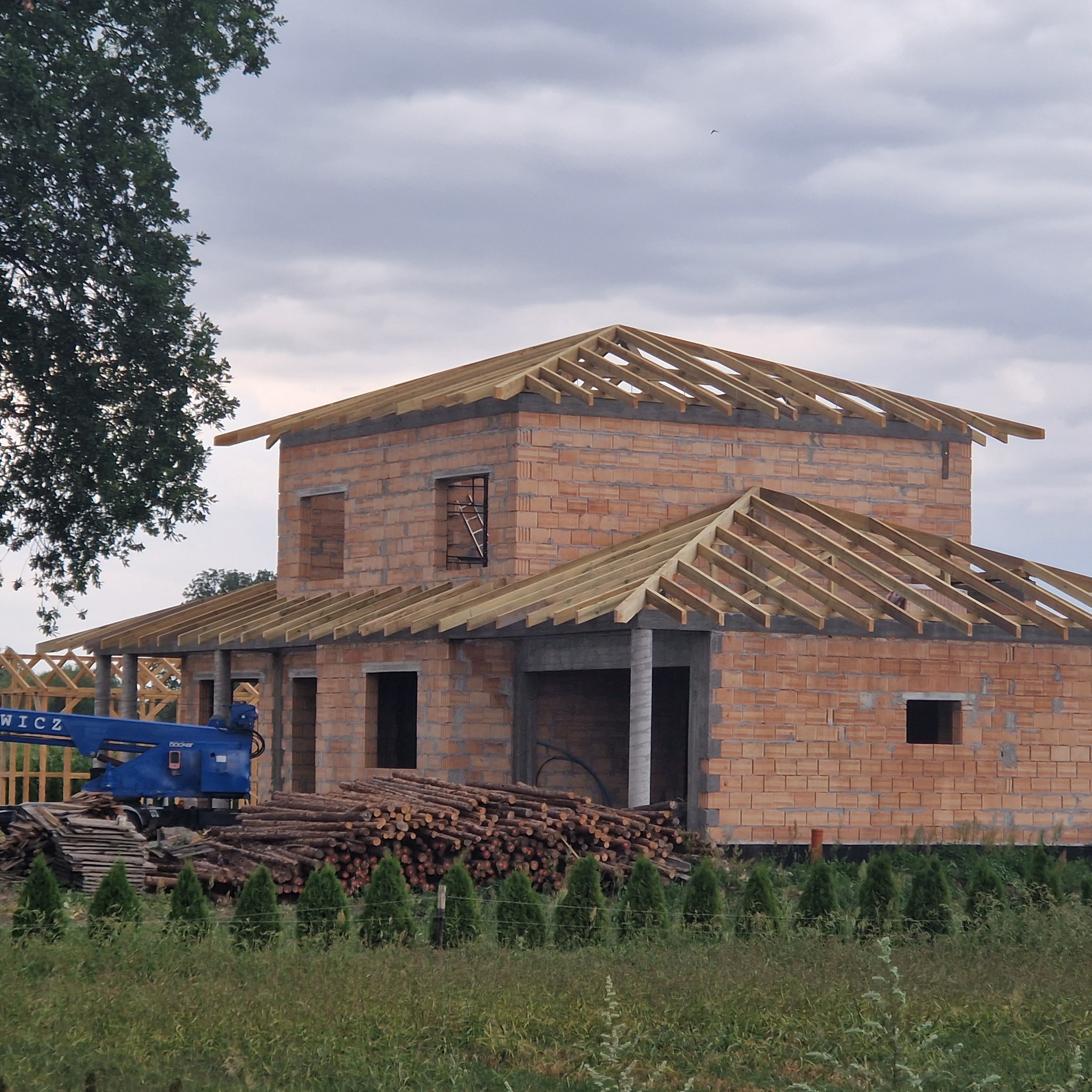 Surowy stan budynku z cegły z drewnianą konstrukcją dachu w trakcie budowy, na pierwszym planie stos drewna i podnośnik koszowy.