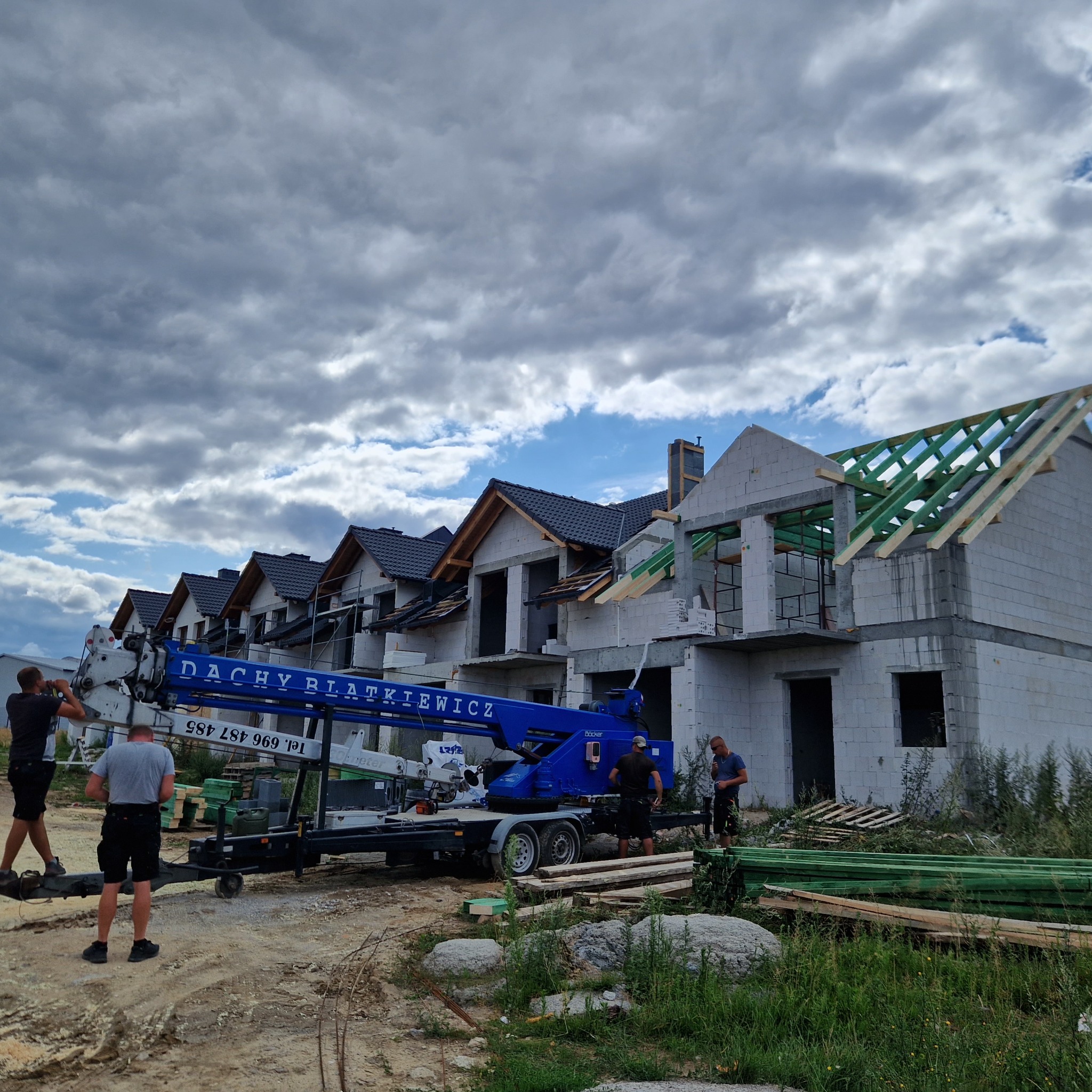 Montaż więźby dachowej na budowie szeregu domów jednorodzinnych z użyciem dźwigu z logo firmy Dachy Błatkiewicz, widoczne elementy konstrukcyjne dachu w kolorze zielonym.