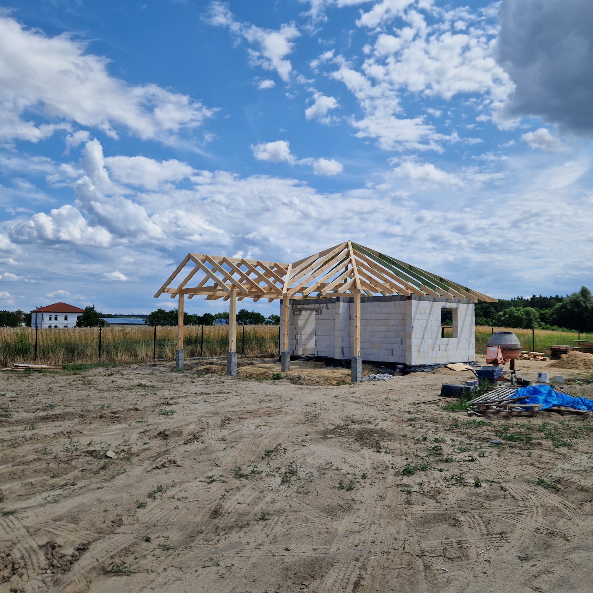 Budowa domu w trakcie realizacji: wzniesione ściany z betonu komórkowego i częściowo wykonana konstrukcja dachu z drewnianych belek, widok na plac budowy w słoneczny dzień.