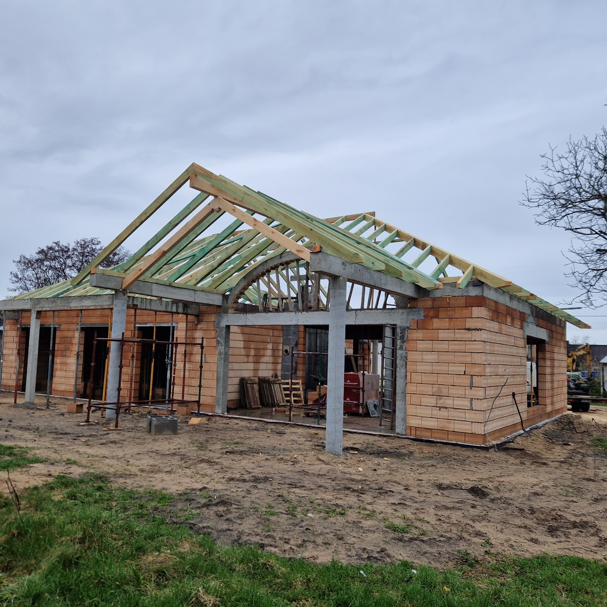Budowa domu jednorodzinnego: surowa konstrukcja z czerwonej cegły, z częściowo zamontowaną drewnianą konstrukcją dachu, widoczne szare betonowe słupy i podciągi, rusztowania w oknach, ziemia wokół...