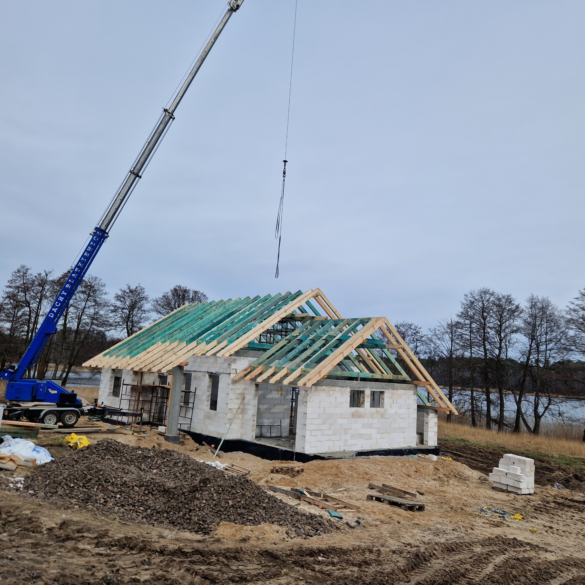 Budowa domu jednorodzinnego w stanie surowym otwartym z użyciem dźwigu, widoczne elementy konstrukcji dachu zaimpregnowane na zielono.