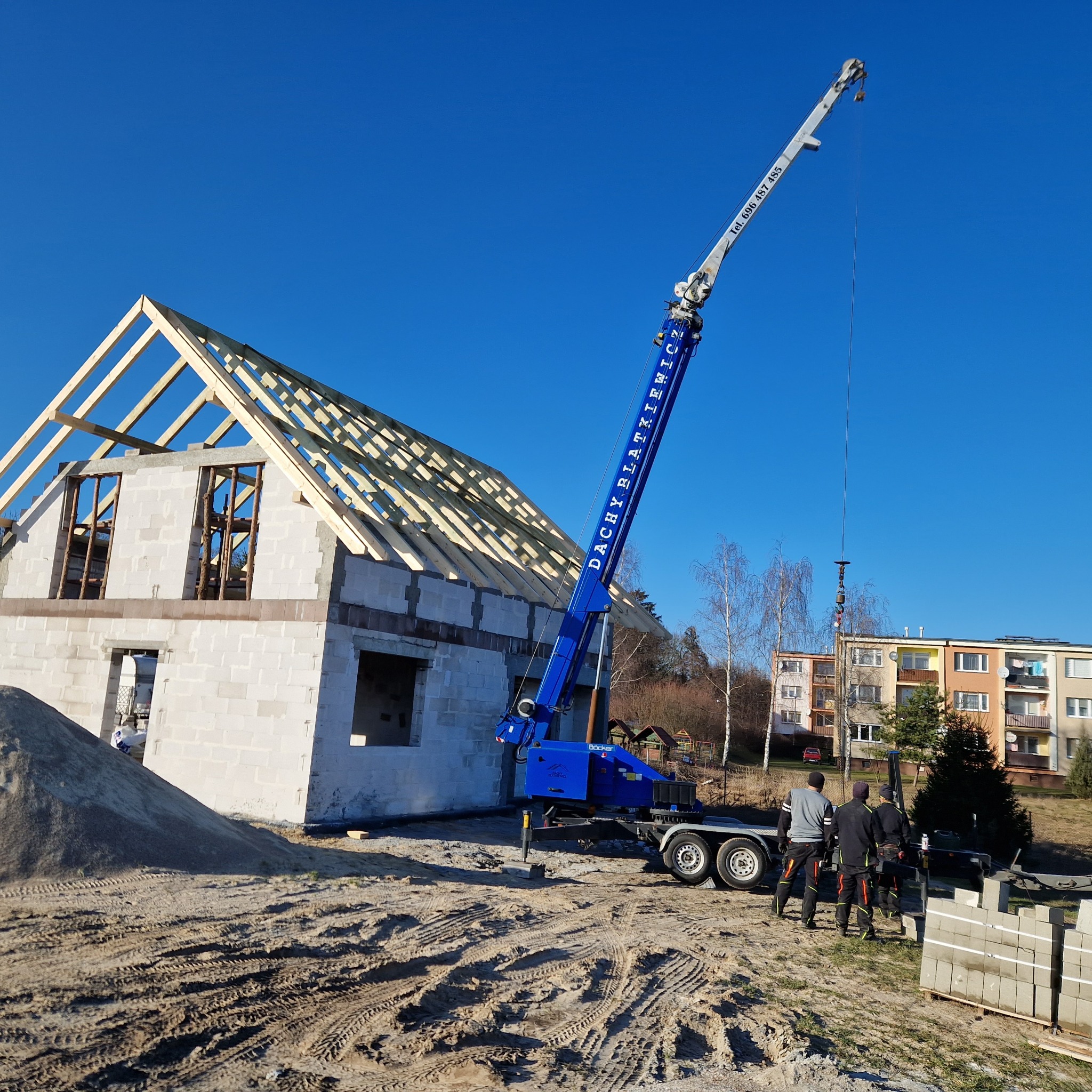 Montaż drewnianej konstrukcji dachu na surowym budynku z pomocą niebieskiego dźwigu z napisem 'DACHY BŁAŻKIEWICZ', w tle pracownicy i budynki mieszkalne.