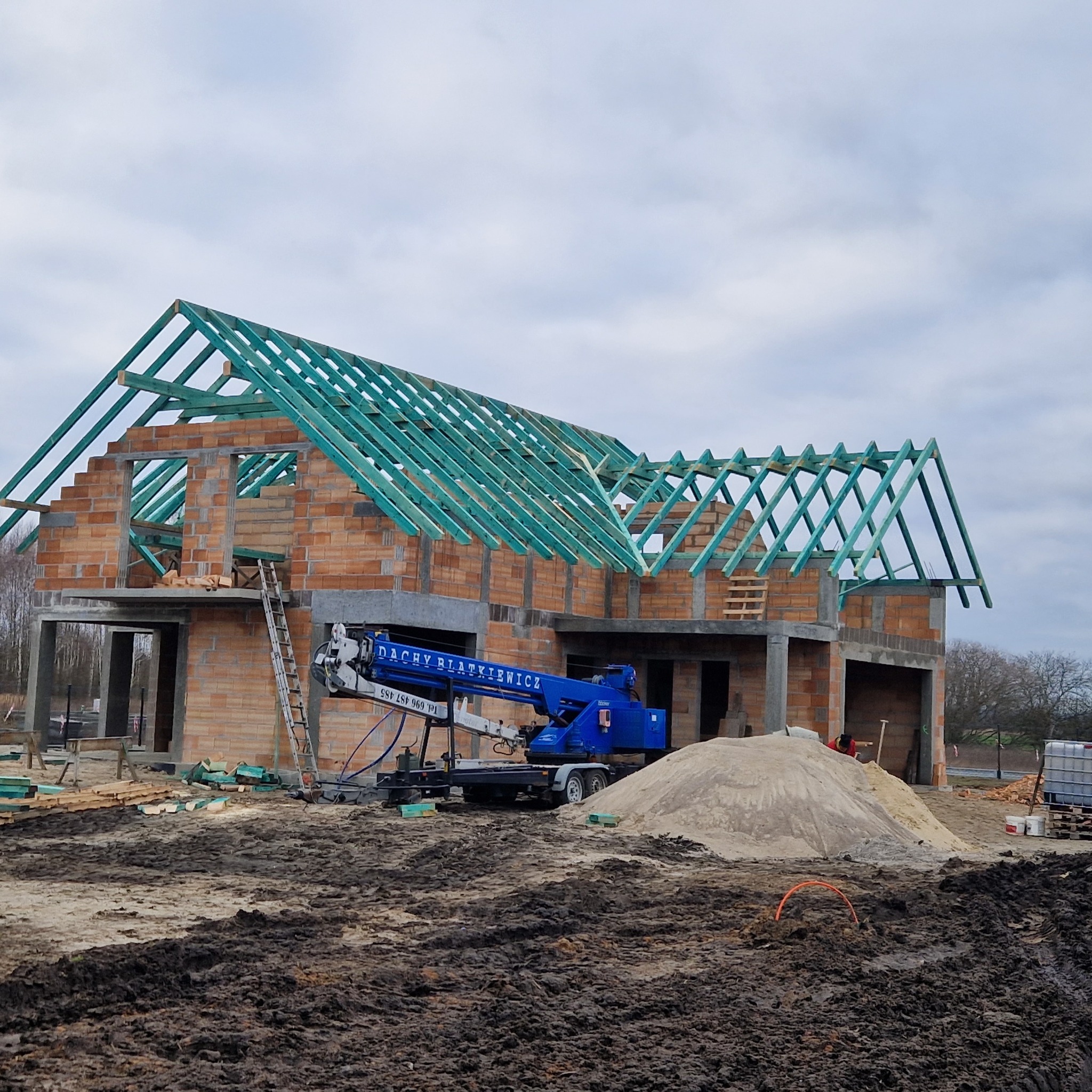 Wznoszenie konstrukcji dachu z impregnowanego drewna na budynku w stanie surowym otwartym, widoczny dźwig z nazwą firmy, teren budowy z błotem i stertą piasku.