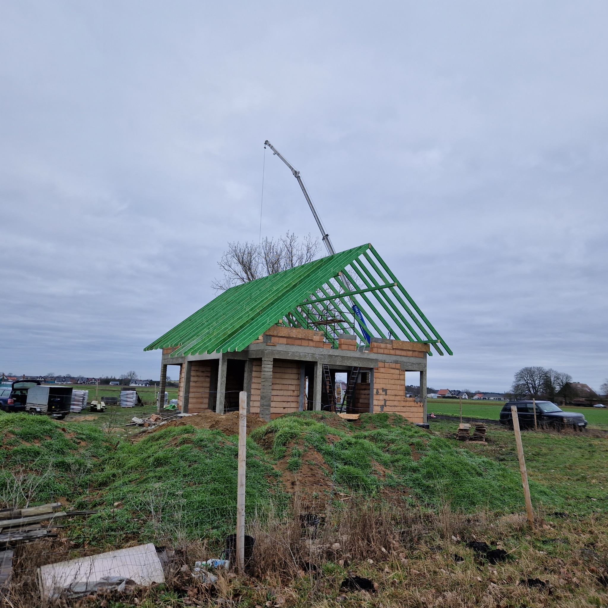 Budowa domu jednorodzinnego w trakcie wznoszenia więźby dachowej z zielonych łat, widoczny dźwig i materiały budowlane na działce.