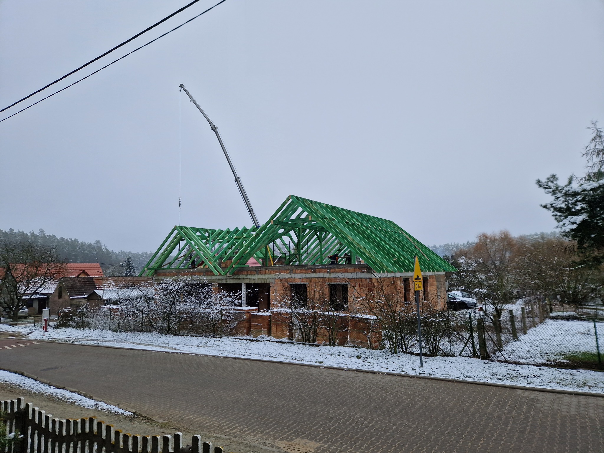 Montaż zielonej konstrukcji dachowej na murowanym domu w zimowej scenerii, widoczny żuraw budowlany i pracownicy na dachu.