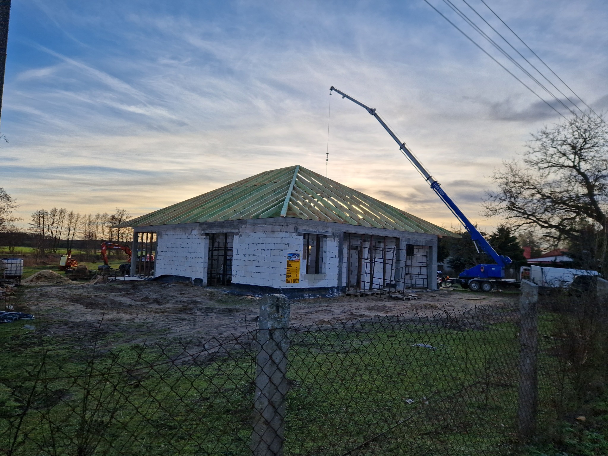 Budowa domu jednorodzinnego w stanie surowym otwartym, widoczna konstrukcja dachu z drewnianych belek, ściany z białego bloczka, na placu budowy dźwig samochodowy i maszyny budowlane, ogrodzenie...