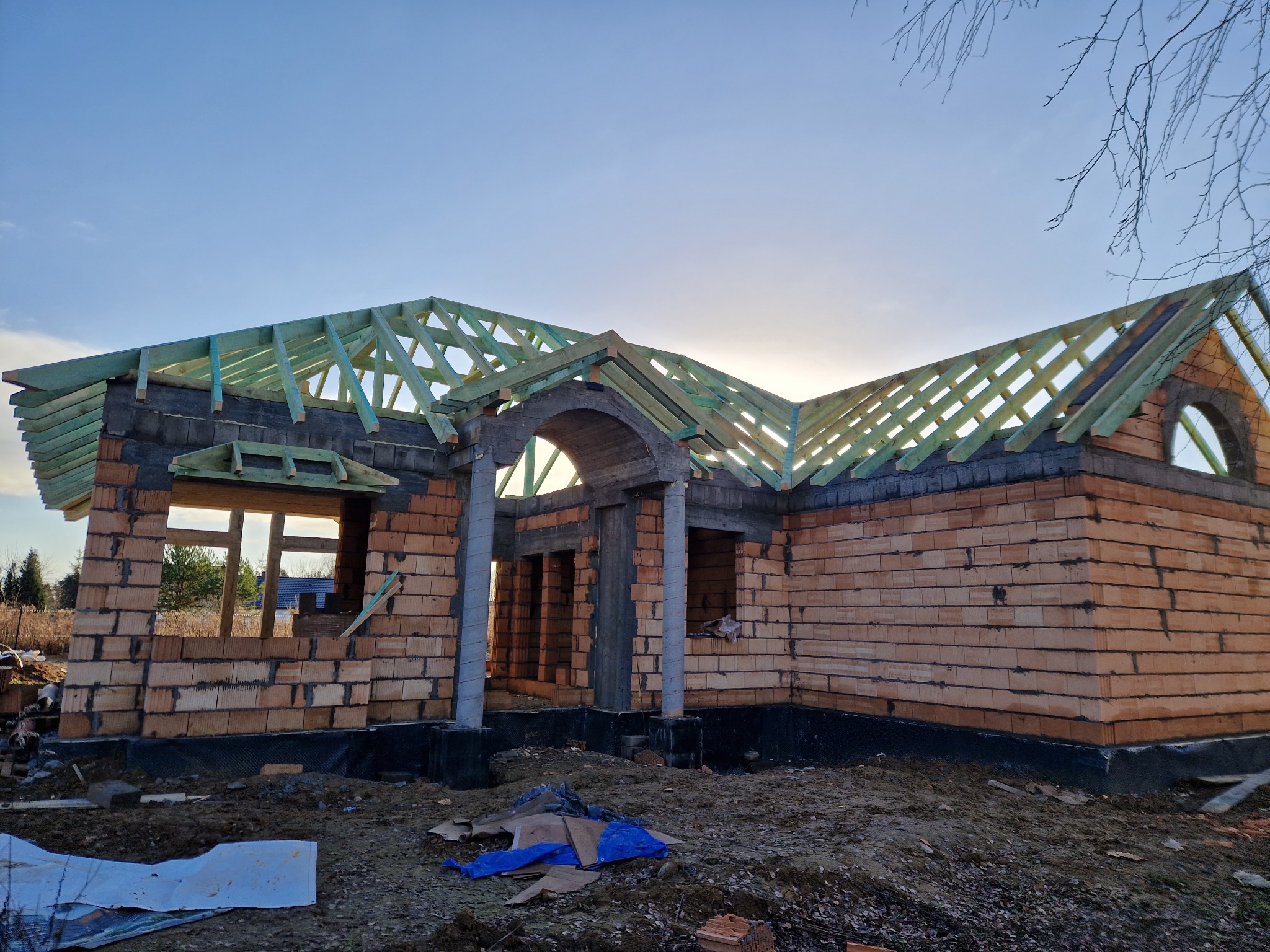 Surowy stan domu jednorodzinnego w trakcie budowy, widoczna konstrukcja dachu z impregnowanego drewna, ściany z cegły ceramicznej, okrągłe okno w szczycie.