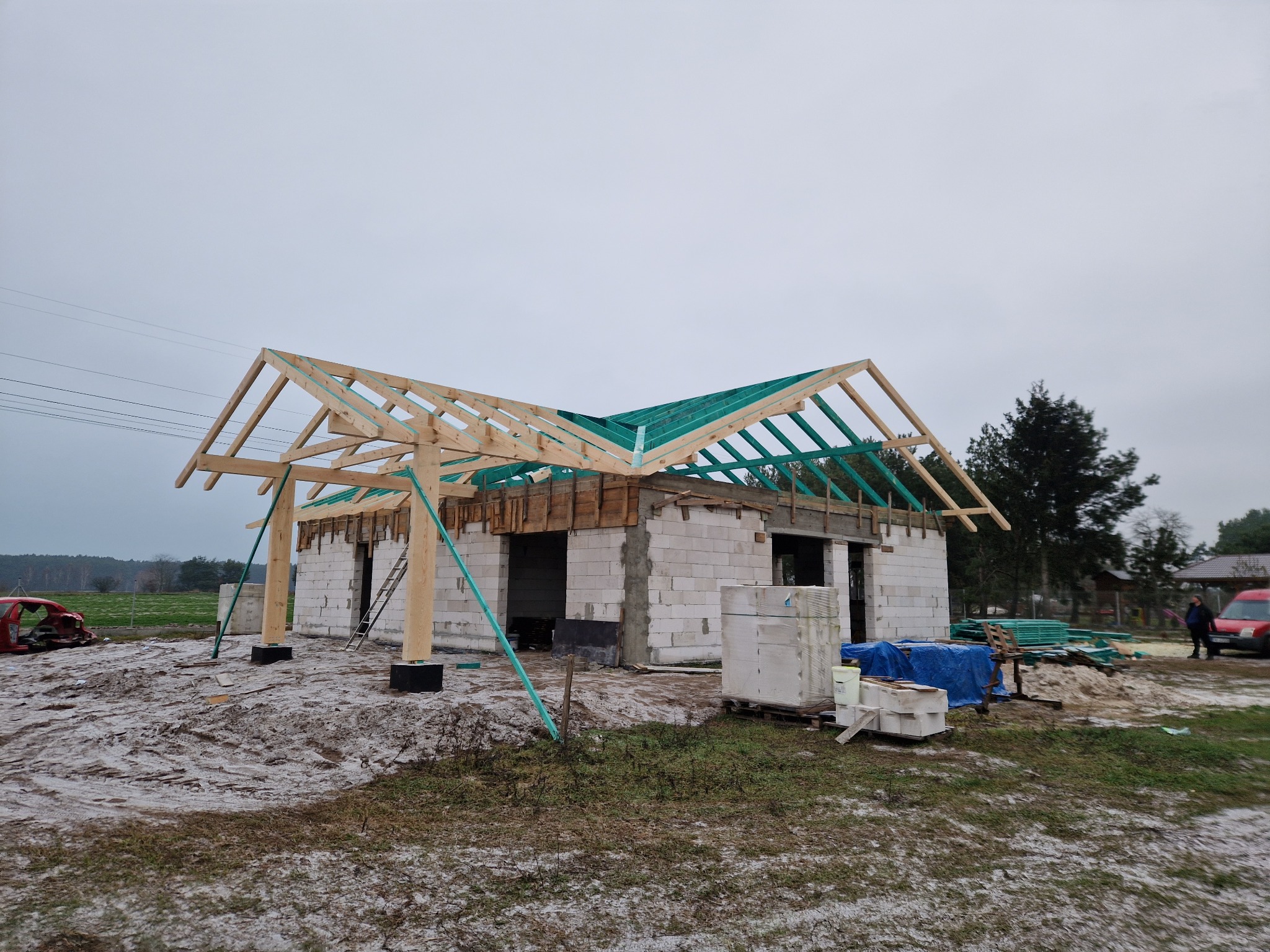 Budowa domu jednorodzinnego w stanie surowym otwartym, widoczna konstrukcja drewniana dachu z zieloną membraną, ściany z białego bloczka, teren budowy z błotem i śniegiem, w tle las i pochmurne niebo.