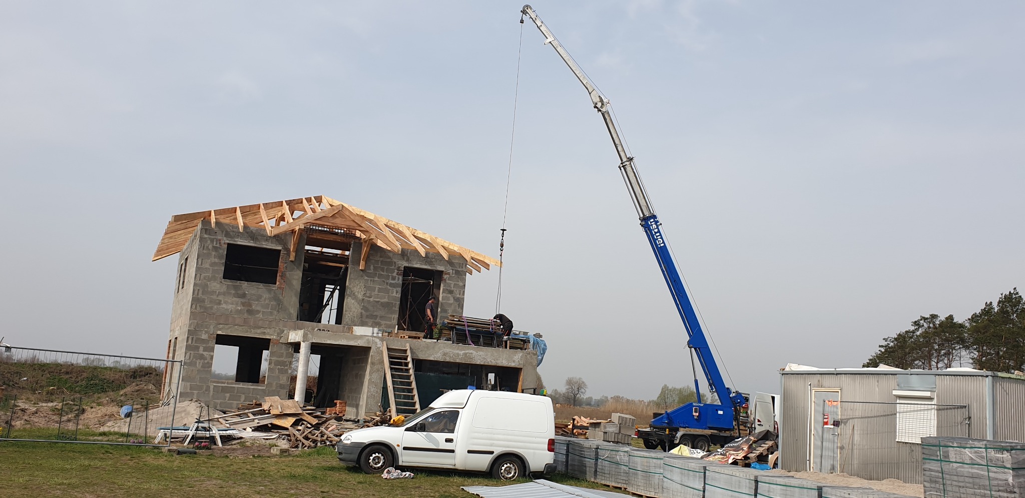 Budowa domu jednorodzinnego w trakcie montażu drewnianej konstrukcji dachu za pomocą dźwigu, widoczny biały samochód dostawczy i materiały budowlane na placu.
