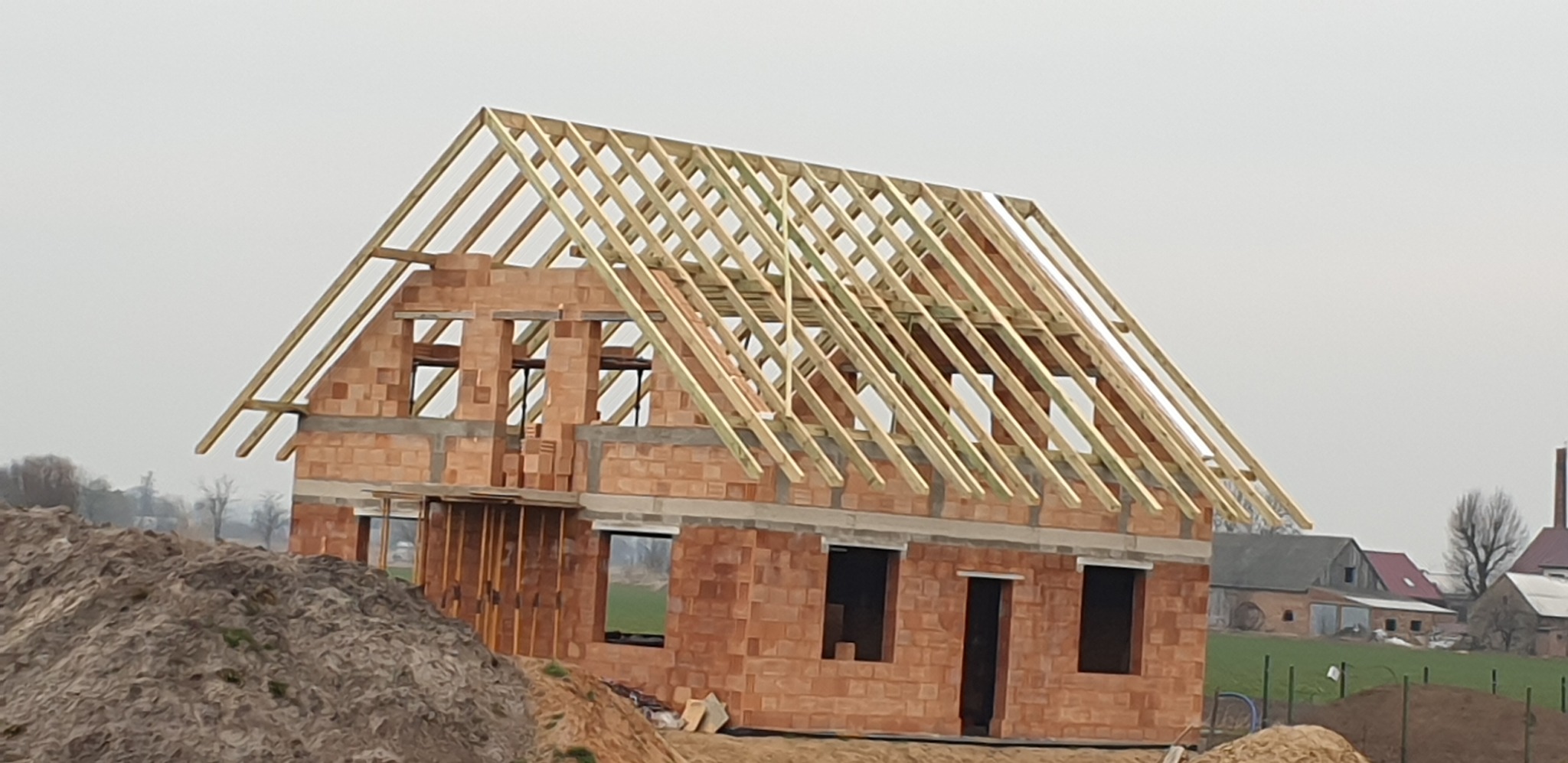 Surowy stan domu z cegły z zamontowaną drewnianą więźbą dachową, widok z poziomu gruntu na tle pochmurnego nieba i zabudowań gospodarczych w oddali.