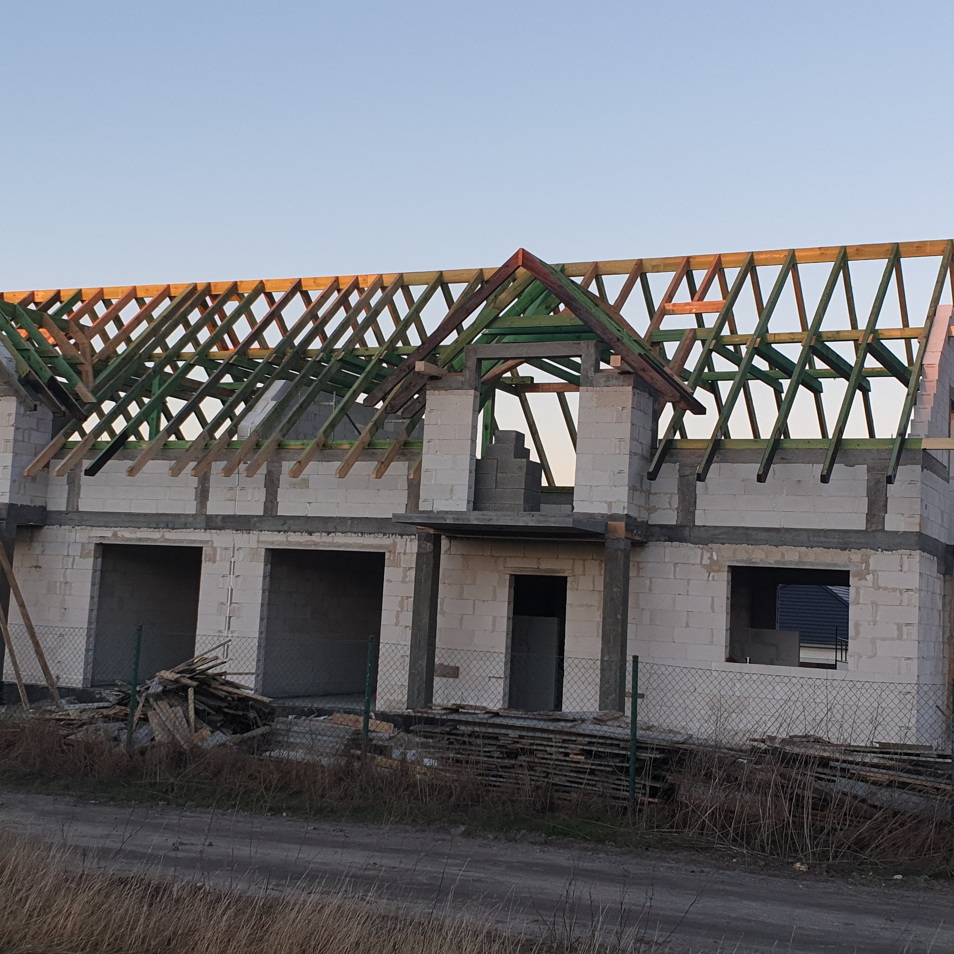 Surowy stan budynku w trakcie budowy, z widoczną konstrukcją drewnianą dachu w kolorze zielonym i brązowym, wykonaną na murach z białego bloczka. Na pierwszym planie widoczny stos drewnianych...