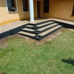 Wykończone schody z kostki brukowej z czarną obwódką prowadzące do domu z żółtą elewacją. Widok z poziomu trawnika.