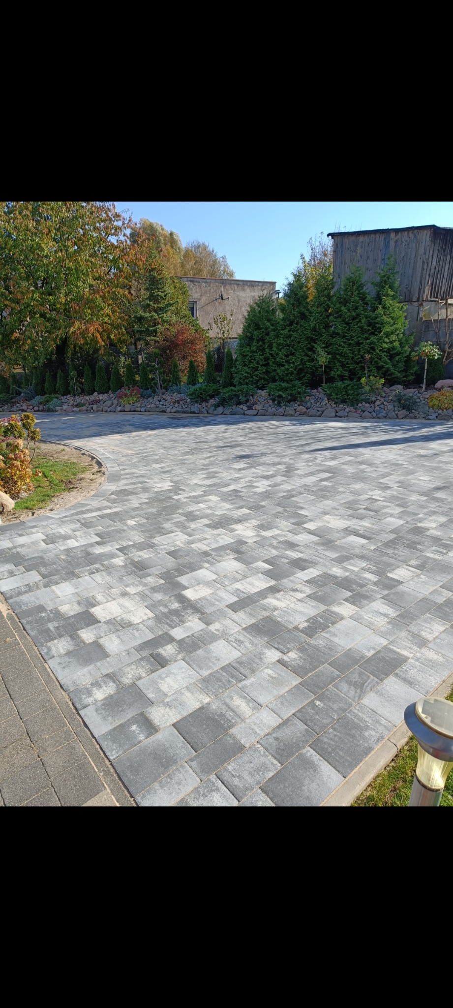 Szara kostka brukowa ułożona na podjeździe, z widocznym fragmentem ogrodu i budynkiem w tle. Oświetlenie solarne wzdłuż krawędzi. Jesienna sceneria.