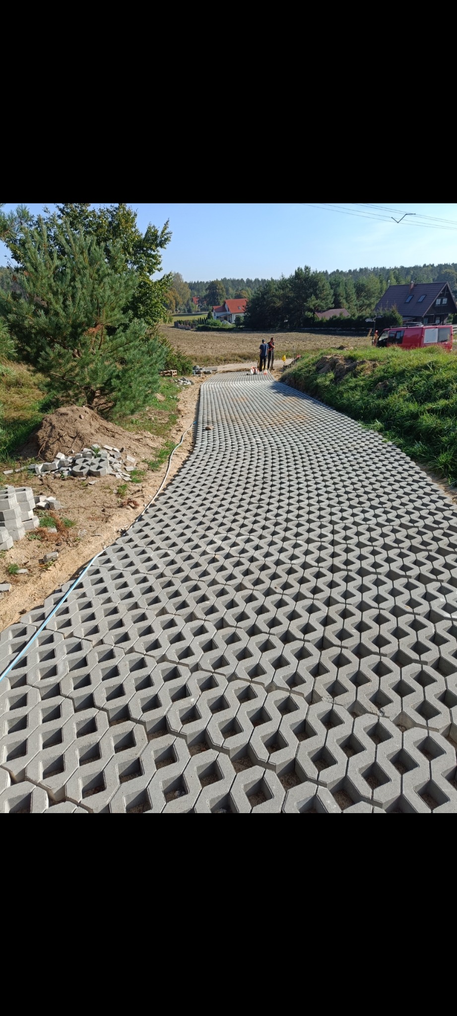Układanie przepuszczalnej kostki brukowej na ścieżce. Widoczne ułożone fragmenty i stos kostki obok. W tle drzewa i zabudowania.