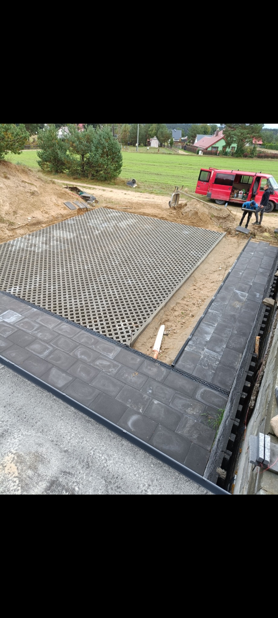 Układanie ażurowych płyt i ciemnej kostki brukowej z odpływem liniowym. W tle czerwony bus i dwóch pracowników. Prace w toku, widok z góry.