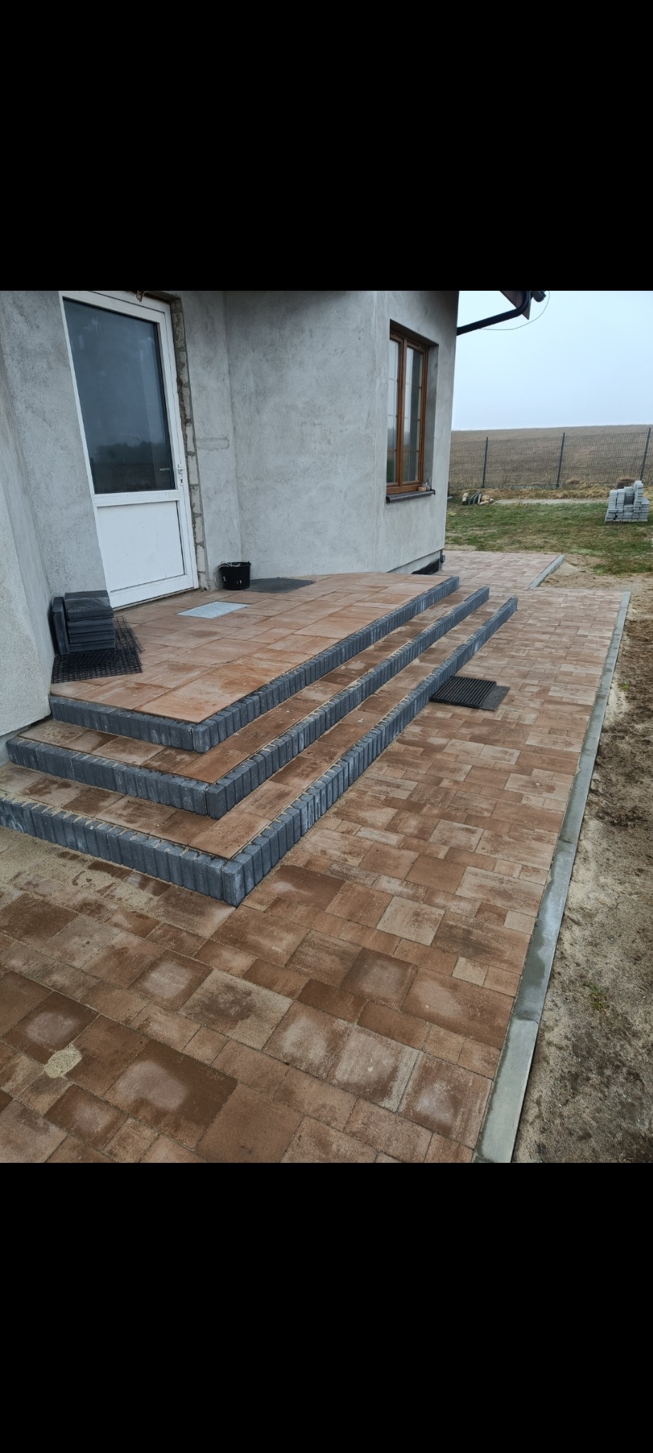 Brązowa kostka brukowa ułożona na schodach i chodniku przed domem. Szare obrzeża kostki. Widoczny fragment elewacji budynku i niezabudowany teren.
