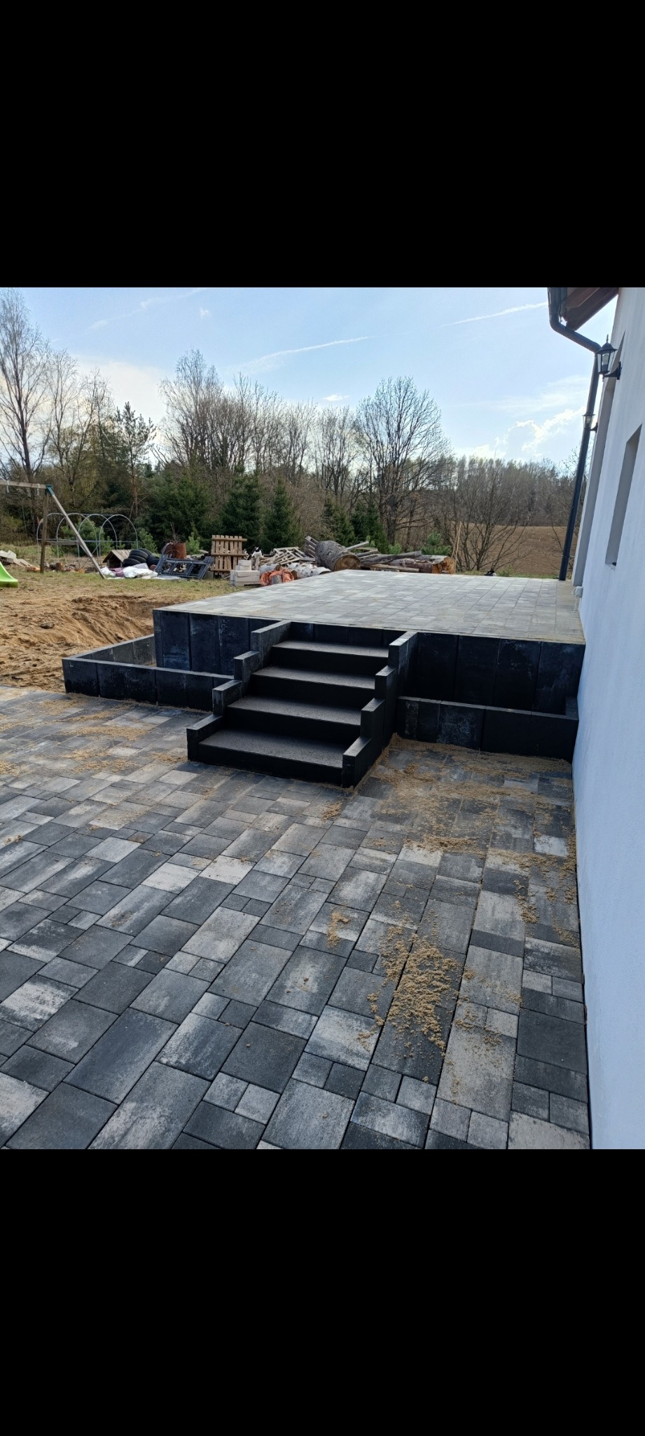 Ułożona, szara kostka brukowa z czarnymi schodami i podwyższeniem tarasu, widok na plac zabaw w tle. Realizacja z dbałością o detale.