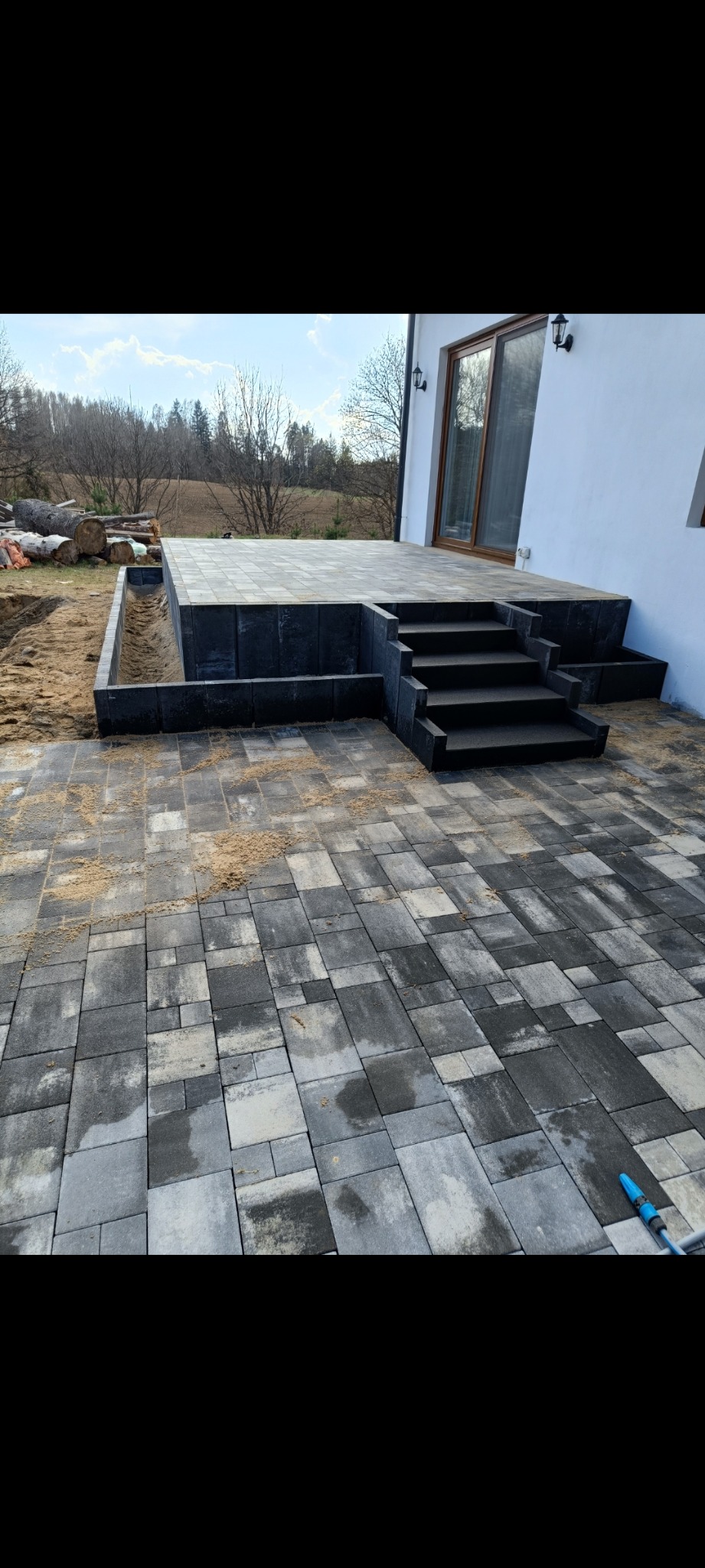 Ułożona, szara kostka brukowa prowadząca do schodów i tarasu przy białym budynku. Widoczne nierówności i piasek na kostce. Częściowo wykończony teren.