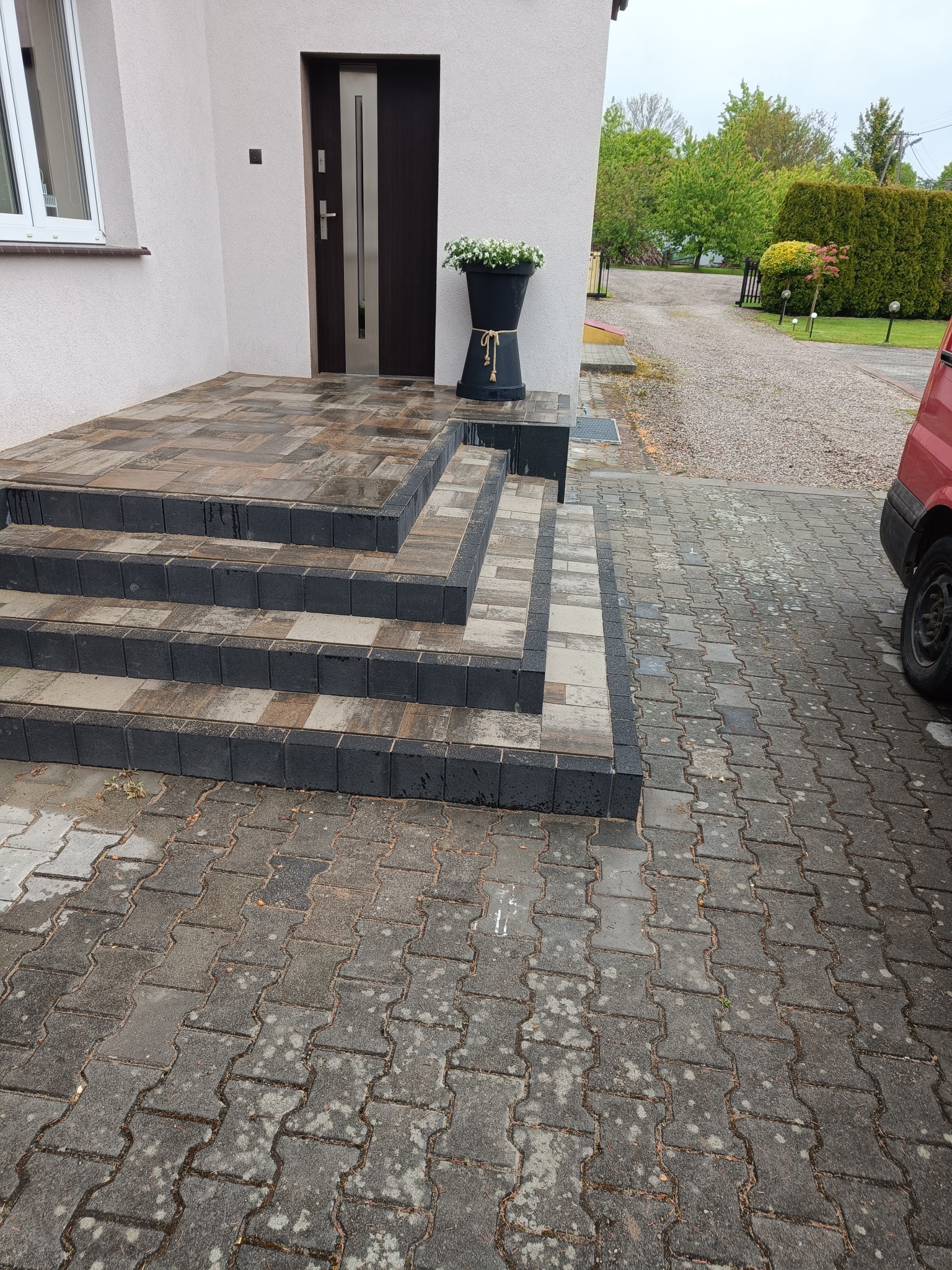 Schody wejściowe z kostki brukowej w odcieniach szarości i brązu, z donicą z kwiatami przy drzwiach. Ułożona kostka prowadzi do żwirowej drogi.