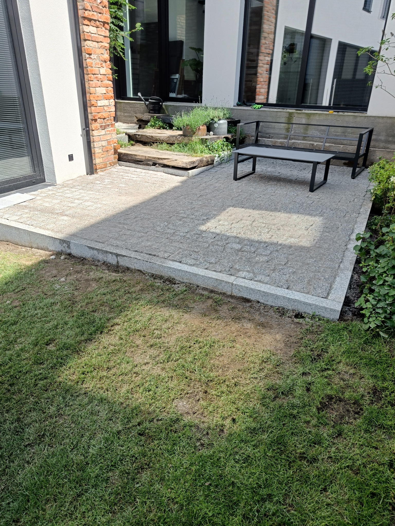 Wykończony taras z kostki granitowej, z szarą obwódką, przylegający do budynku z ceglanym elementem. Na tarasie stoi metalowa ławka z blatem.