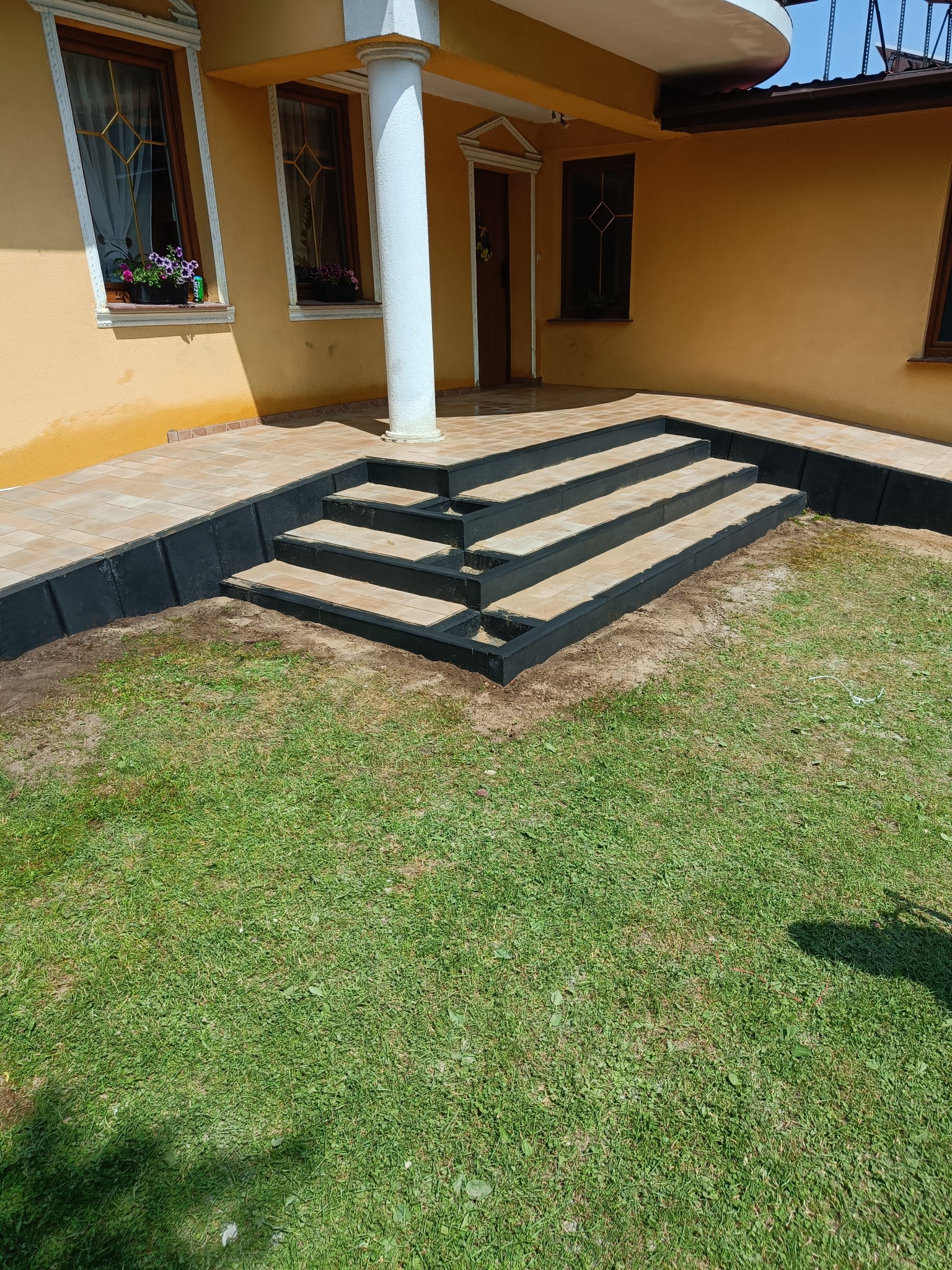 Wykończone schody z kostki brukowej z czarną obwódką prowadzące do domu z żółtą elewacją. Widok z poziomu trawnika.