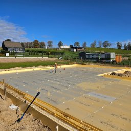 Prowood Sławomir Szczukowski - Fundament domu z izolacją XPS na tle zielonego krajobrazu i błękitnego nieba. Widoczny niwelator laserowy, co sugeruje precyzyjne przygotowanie podłoża.