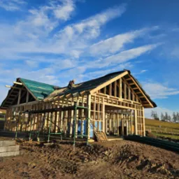 Konstrukcja domu szkieletowego w trakcie budowy z widocznym pracownikiem na dachu, błękitne niebo w tle. Elementy szkieletu drewnianego.