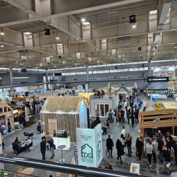 Prowood Sławomir Szczukowski - Widok z góry na halę targową z ekspozycjami domów modułowych i szkieletowych, liczne osoby zwiedzają stoiska. Jasne oświetlenie, widoczne konstrukcje dachowe hali.