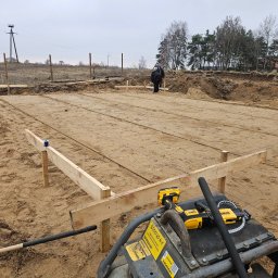 Prowood Sławomir Szczukowski - Przygotowany teren pod budowę domu: wytyczone fundamenty, drewniane szalunki i sprzęt budowlany na piaszczystym podłożu. Widoczni pracownicy w tle.