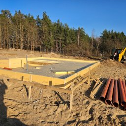 Prowood Sławomir Szczukowski - Fundament domu z izolacją XPS, przygotowany pod budowę. Na placu budowy widoczny żółty traktor i rury kanalizacyjne. W tle las i błękitne niebo, cień osoby w lewym rogu.