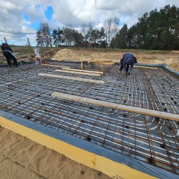 Prowood Sławomir Szczukowski - Montaż ogrzewania podłogowego w fundamencie domu szkieletowego. Widoczne rury grzewcze, siatka zbrojeniowa i pracownicy. Fundament zabezpieczony izolacją.