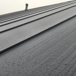 Prowood Sławomir Szczukowski - Szary, blaszany dach z widocznymi kroplami deszczu i kominkiem w tle, sfotografowany pod kątem. Widoczne regularne linie blachy dachowej.