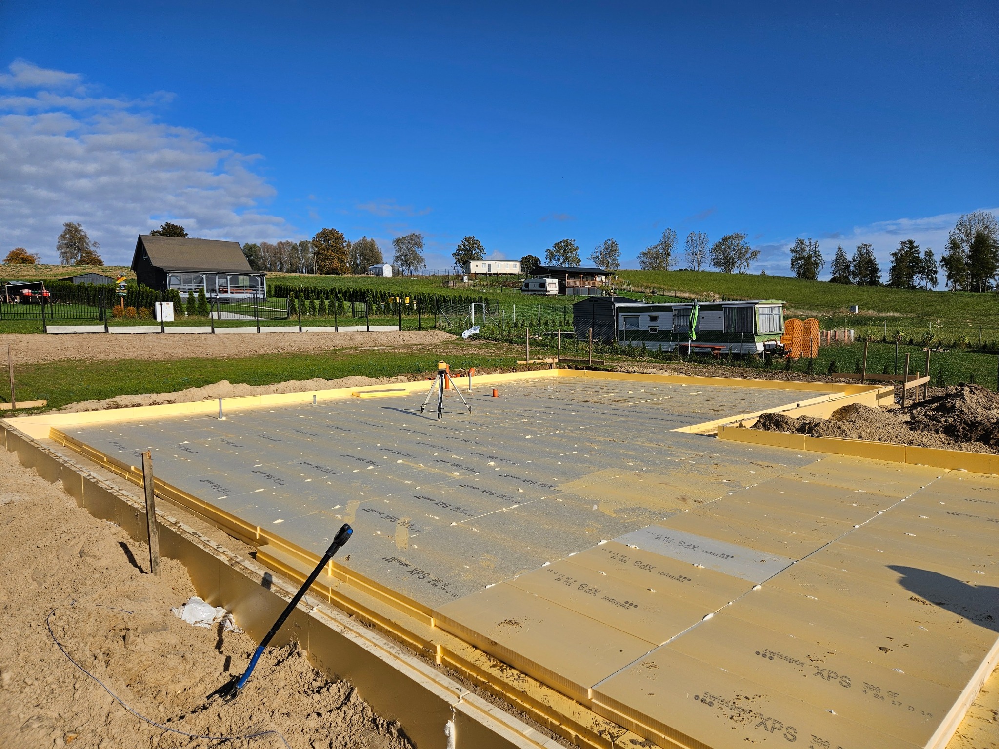 Fundament domu z izolacją XPS na tle zielonego krajobrazu i błękitnego nieba. Widoczny niwelator laserowy, co sugeruje precyzyjne przygotowanie podłoża.