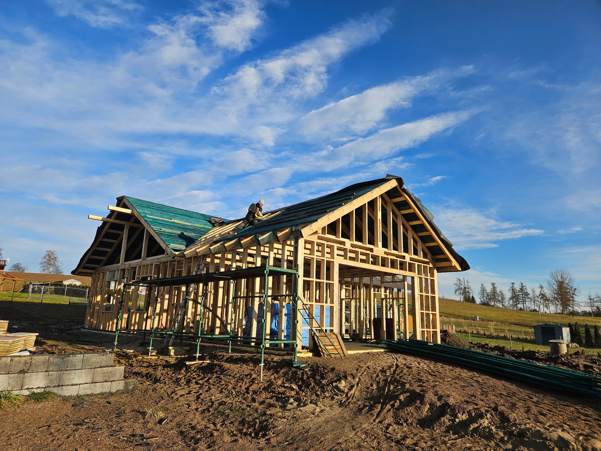Konstrukcja domu szkieletowego w trakcie budowy z widocznym pracownikiem na dachu, błękitne niebo w tle. Elementy szkieletu drewnianego.