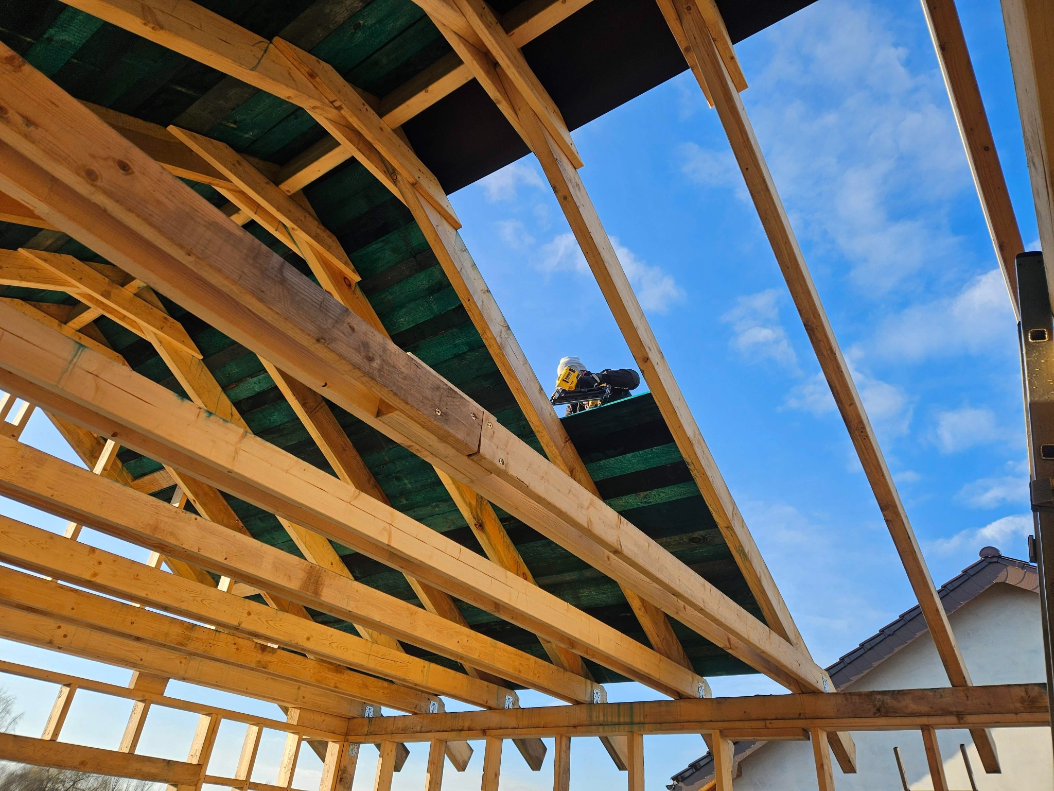 Wnętrze konstrukcji drewnianej domu w budowie z widocznym niebieskim niebem w tle, z użyciem narzędzi do montażu. Widok z dołu na szkielet dachu.