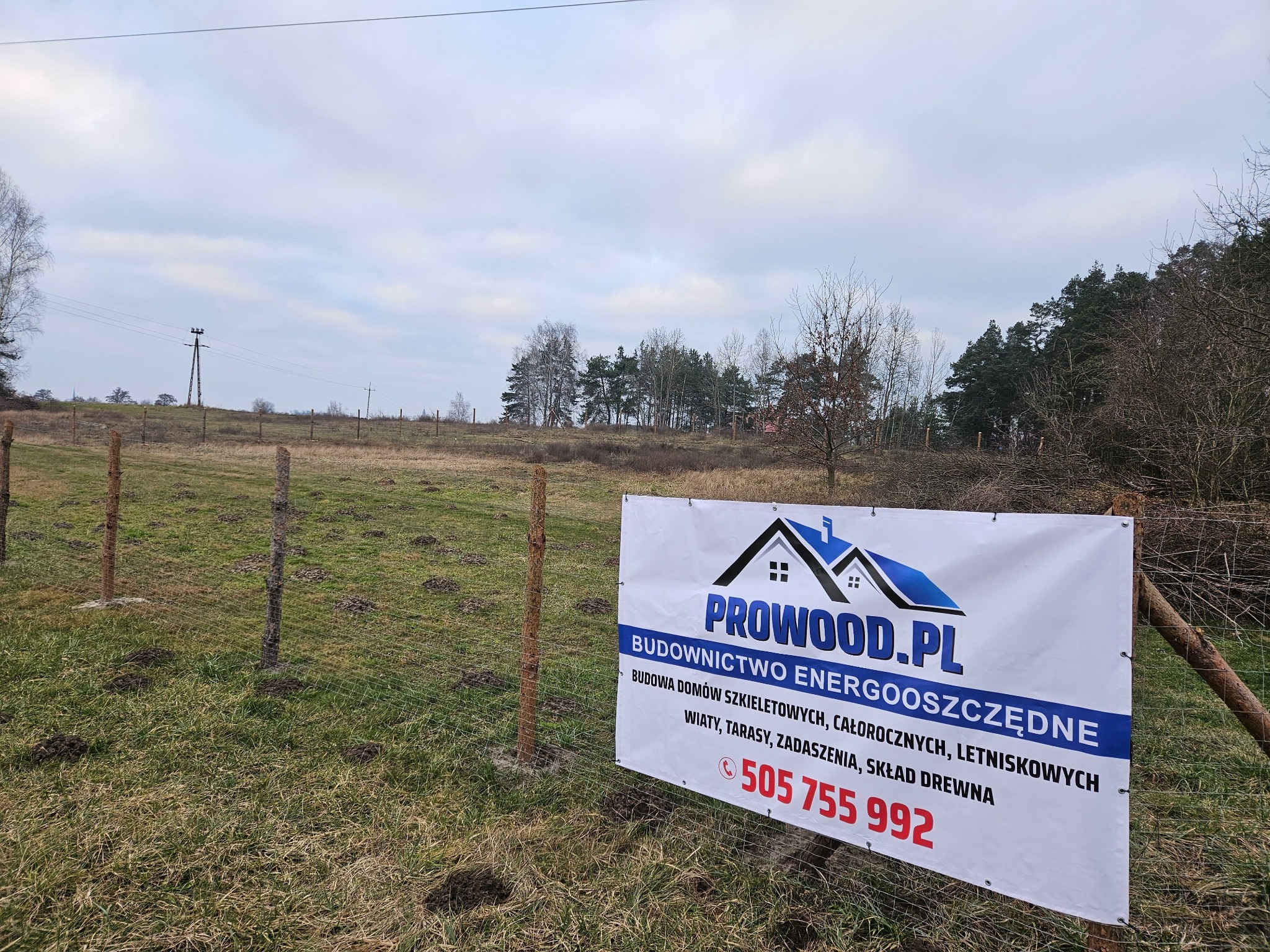 Baner firmy Prowood.pl na tle pola z ogrodzeniem. Oferta: budowa domów szkieletowych, całorocznych, letniskowych, wiaty, tarasy, zadaszenia, skład drewna. Numer telefonu.